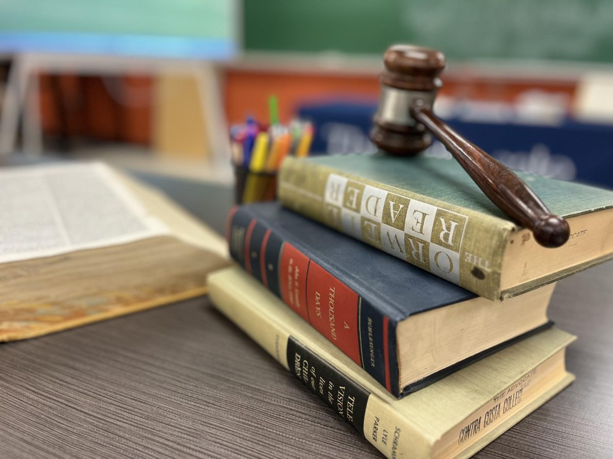 Books and a gavel sit on a classroom desk.The image reflects the tension in education debates: the tools of knowledge and the authority to judge them.
